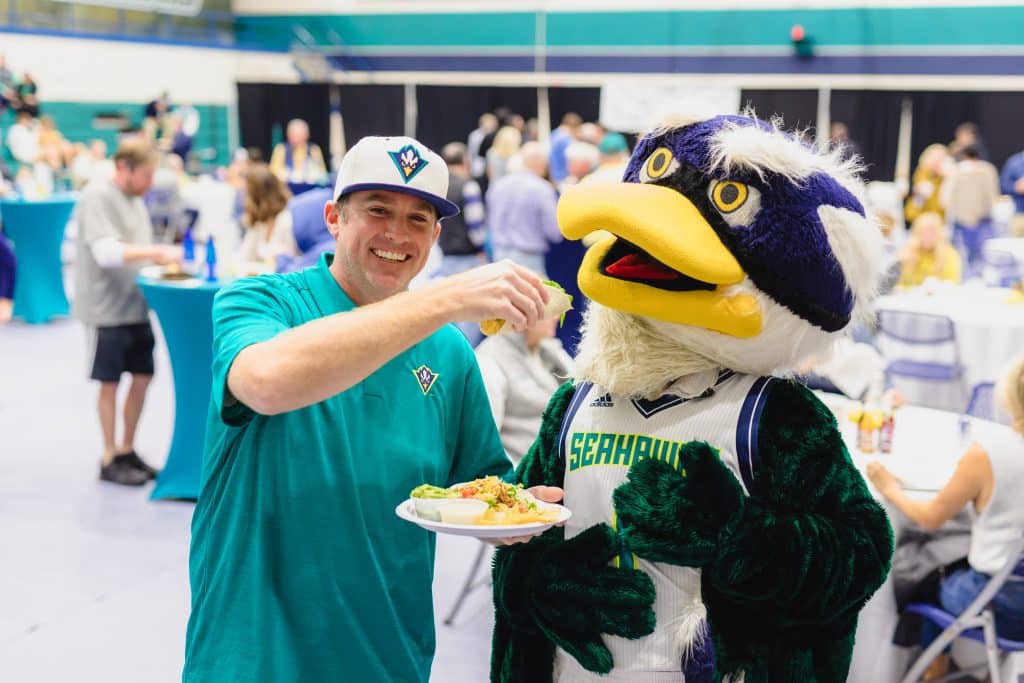 A man wearing a green shirt and blue hat is holding a plate of food, smiling at the camera.