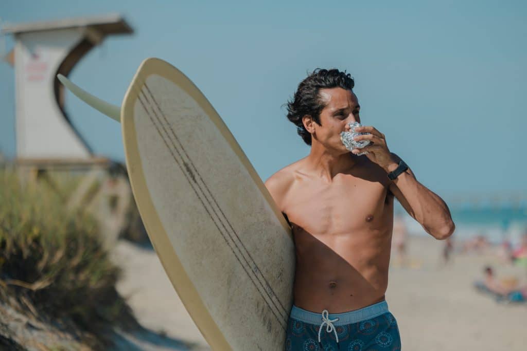 A man in swimming shorts holding a surfboard and a wrap standing on a beach.